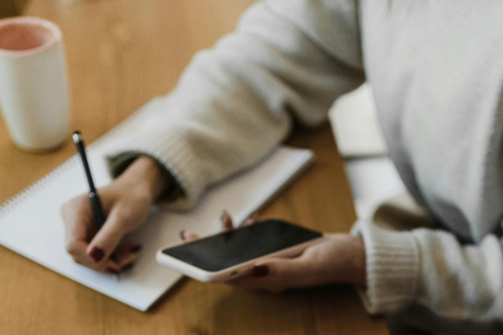 A person in a white sweater consults a mobile phone while writing on a notepad.